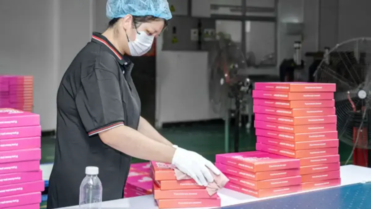 A gloved worker wiping and stacking finished card boxes at a packing table in a factory