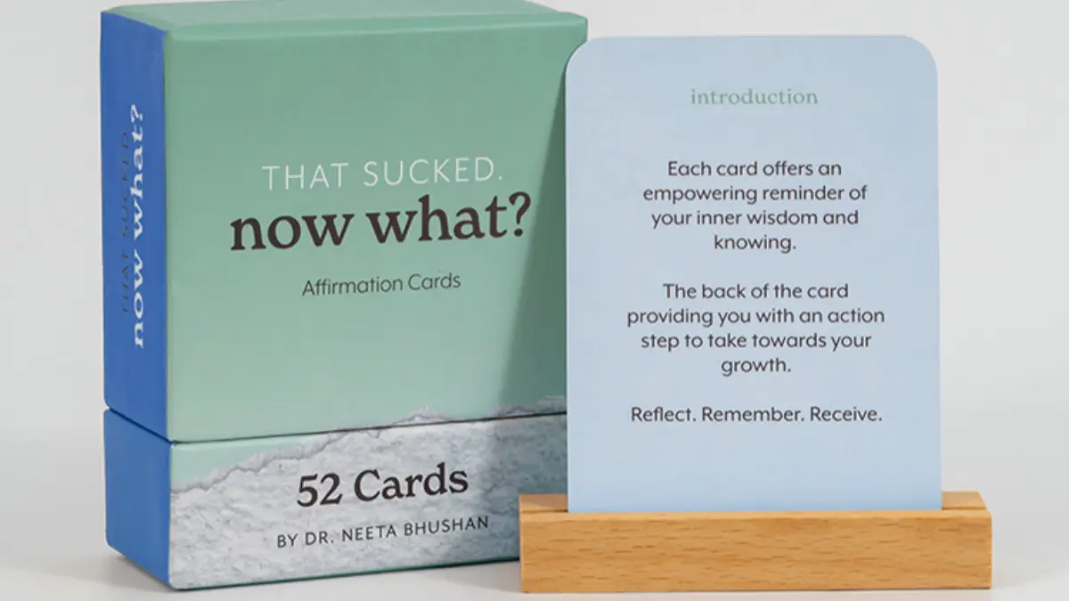 Teal and blue affirmation card box labelled That Sucked now what beside a 52-card deck and a card held on a wooden stand